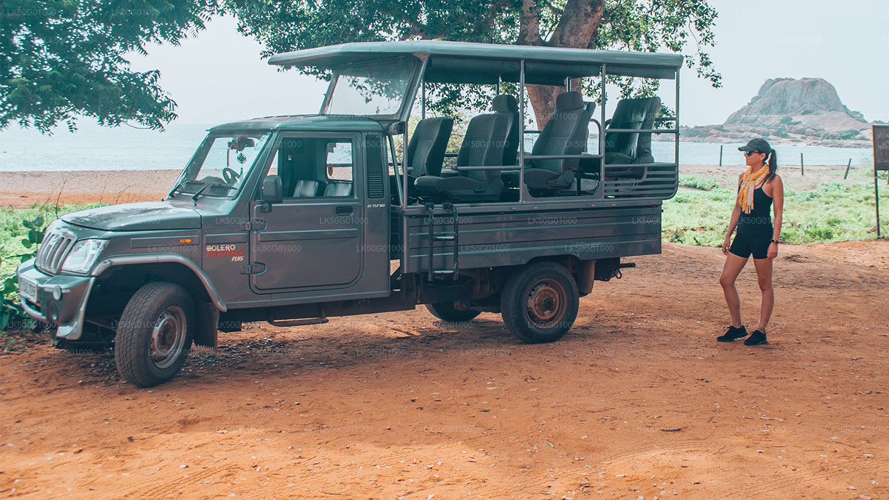 Elephant safari jeep parked under a tree with a woman standing nearby, set against a backdrop of sandy ground, sea, and distant rocky landscape.
