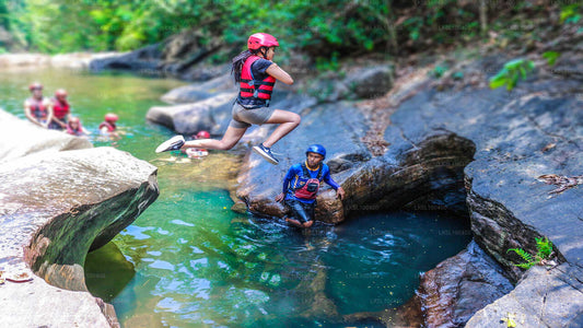 A group of people canyoning and rafting in a natural water setting with rocks and trees in the background. One person is jumping off a rock into the water, while others watch and prepare to descend.
